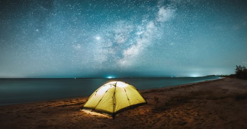 Starry blue and white sky shining bright at night above yellow tent alongside a beach.