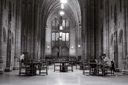 A black-and-white shot of the interior of the Cathedral of Learning building at the University of Pittsburgh in Pittsburgh, Pennsylvania.There are several small tables with chairs around them. A student with headphones is sitting at one of the tables, looking at a laptop computer.