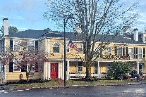 An exterior view of the historic Concord's Colonial Inn in Concord, Massachusetts. The inn is yellow and has a red door. An American flag on a flagpole flies in front of the inn.