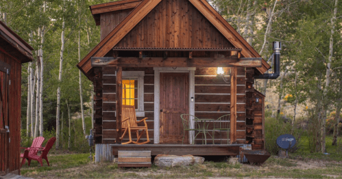 A log cabin with a rocking chair on the porch stands in front of a copse of trees.