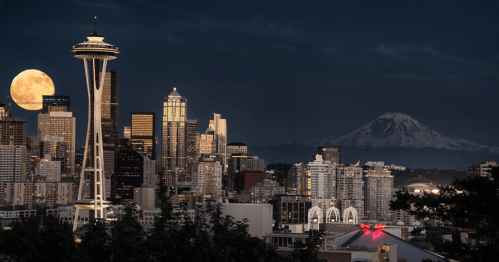 The Seattle skyline sparkles beneath a full moon at night time.