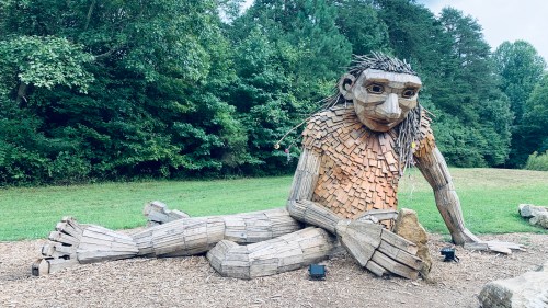 A wooden troll statue lounges along the ground while playing with rocks.