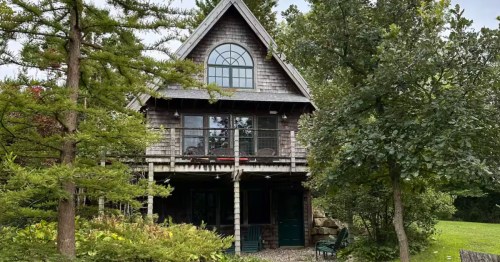 Charming two-story Eagle Cozy Cottage exterior. Wooden cabin surrounded by lush trees, featuring an arched window and balcony.