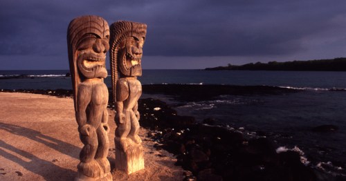 Wooden kiʻi statues and thatched structures at Puʻuhonua o Hōnaunau National Historical Park on the Big Island.