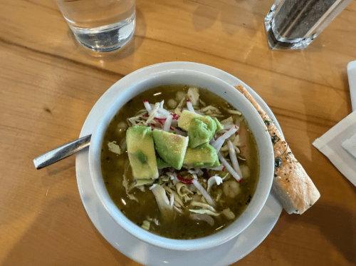 Fresh chunks of avocado and shredded cabbage top soup in a white bowl on a white plate on a brown wooden table. A slice of focaccia sits on the plate next to the bowl.