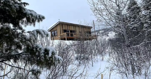 A small cabin with wood-planked walls and a patio on one side sits on top of a snowy hill with barren and snow-covered trees surrounding it.