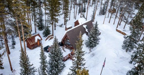 A modern A-frame cabin and a small outbuilding surrounded by snow-covered pine trees in a dense Colorado forest.