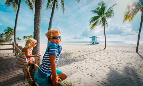 A joyful young boy and girl standing side by side on a tropical beach, both smiling brightly with wind-tousled hair, wearing casual summer clothes