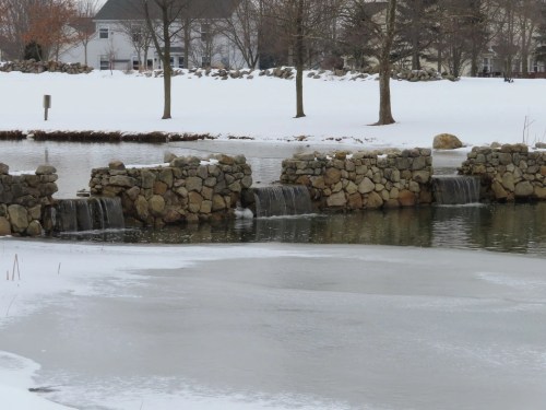 Three man-made waterfalls trickle into a flowing river despite snow-covered banks.