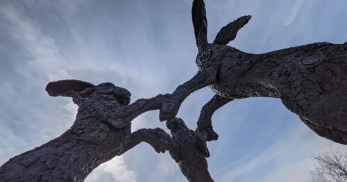A bronze statue featuring three dancing bunnies with outstretched and interlocked arms is shadowed against a blue cloudy sky.
