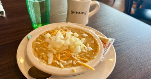 A bowl of chicken chili topped with onions and shredded cheese sits on a plate near a white coffee mug that reads "yummy."