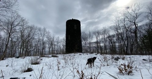A dark, round stone tower stands on a snow-covered hill surrounded by bare trees under a heavily clouded sky, with a dog walking in the foreground.