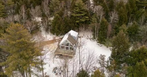 An aerial view shows a modern A-frame cabin with large windows nestled in a snowy, evergreen forest.