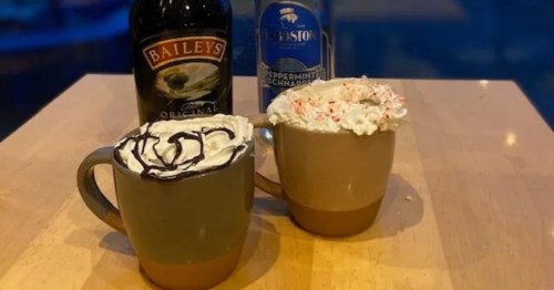 Two festive mugs of hot chocolate with toppings (whipped cream, syrup) are on a wooden table with bottles of liqueur.
