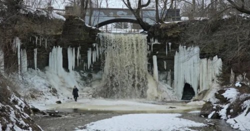 Beneath a snow-covered bridge, a frozen waterfall in Wisconsin trickles down into a still, snow-covered river.