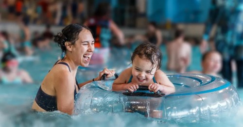 A woman and young child laughing while playing with a clear inflatable tube in a crowded indoor pool.