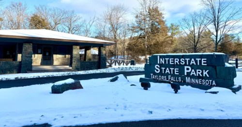 A large stone sign for Interstate State Park in Taylors Falls, Minnesota, sits on a snowy lawn next to a park building.