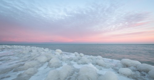 A picturesque winter sunset or sunrise over a large body of water, featuring a foreground of large, white, rounded ice formations and a sky of soft pink and light blue colors.