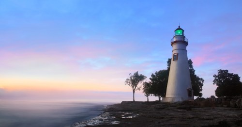 A white lighthouse with a green light stands on a rocky shore beside a body of water at sunrise or sunset.