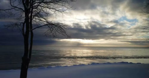 * A peaceful sunrise scene on Whitefish Bay in Paradise, Michigan. The sun is rising over the horizon, casting its light on the snow-covered shore.