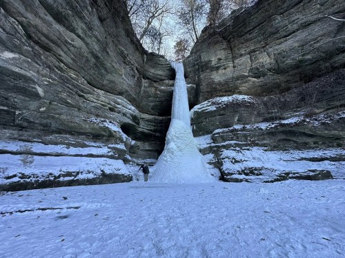 A massive trickle of ice dwarfs a person standing before a stone-covered sandstone cliff in Illinois.