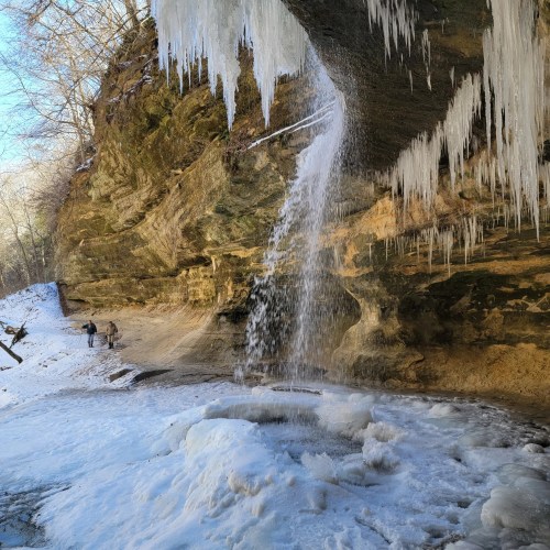 Water cascades off an ice-covered sandstone cliff in Illinois as hikers approach.
