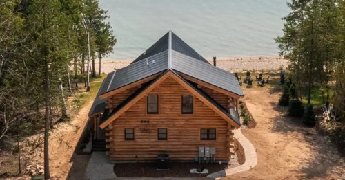 A stunning resort-style cabin rental nestled on the shores of Lake Michigan in Ellsworth, Michigan. The cabin boasts a classic log exterior, contrasting beautifully with the bright blue sky and the sparkling water of the lake. The expansive property offers breathtaking views, with a private beach and a small boat anchored offshore adding to the idyllic scene.