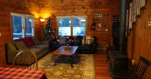 Rustic wood-paneled room with a Christmas tree, wood-burning stove, and plaid tablecloth.