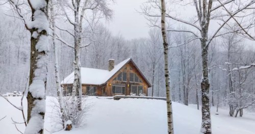 A wood, snow-capped cabin with large windows sits on a snowy hill, surrounded by trees at this Airbnb in Enosburg, Vermont.
