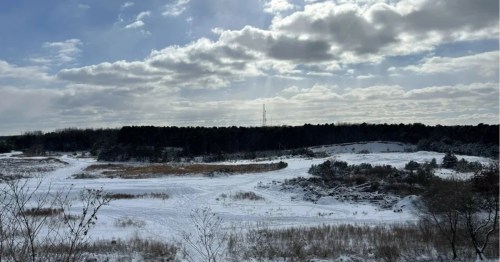 A snow-covered field and frozen wetland area with dark forest in the background under a partly cloudy sky with sun rays.