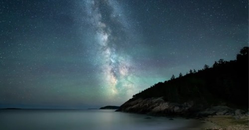 * Milky Way galaxy stretches across a dark sky above a calm ocean and rocky coastline in Acadia National Park.