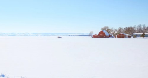A vast snow-covered field leading to a cluster of red farm buildings and bare trees under a bright, pale blue sky.