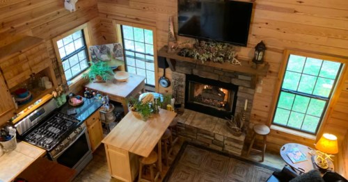 View from the second level of the cabin looking down to the open living room and kitchen. There's natural wood paneling, expansive windows, and a fire going.