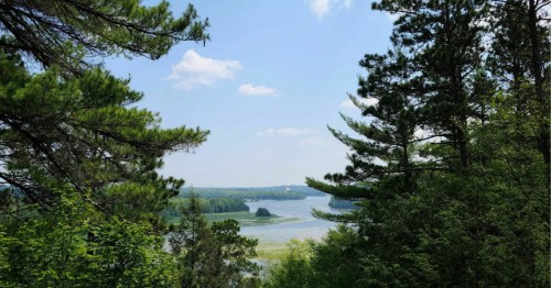 Pine river reflects a blue sky through thick trees, with a tiny village nestled in the lush, distant landscape.