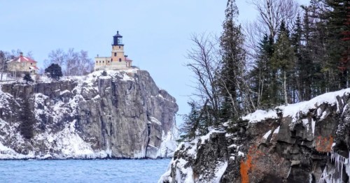Tan lighthouse with black top and red-roofed keeper's house on an ice-covered cliff above Lake Superior's steely blue water.