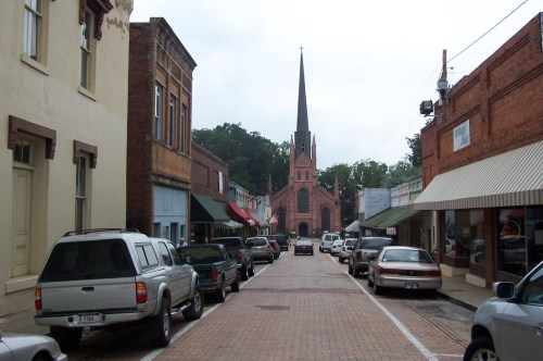 Brick buildings line the brick-covered street ending with a tall Gothic-style church with a looming steeple.