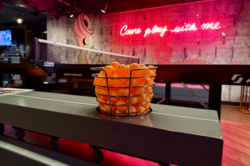 A close-up shot of a bin of orange ping-pong balls in front of a ping-pong table at the SPIN Boston entertainment venue. A neon sign in the background reads "Come play with me."