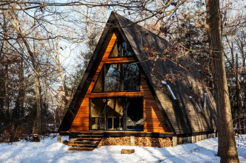 A cozy A-frame cabin with large windows nestled in a snowy forest with a firewood stack underneath.