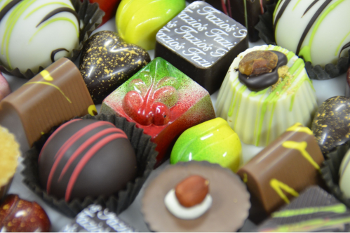 A close-up of assorted Fazio's chocolates in paper cups shows a variety of colors, shapes, and toppings, including swirls, nuts, and stripes.
