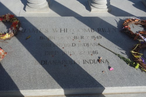 A close-up photograph of the epitaph at the James Whitcomb Riley grave site in Crown Hill Cemetery, Indianapolis, Indiana. The epitaph lists his year of birth and death, both of which occurred in Indiana. Flowers left by visitors drape over the stone, trailing bright colors over a gray background.