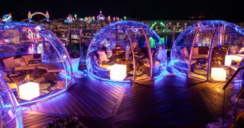 Small groups of people sit inside clear igloos on a wooden deck overlooking Lake Coeur d'Alene at night.