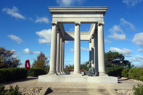 A photograph of the James Whitcomb Riley grave site at Crown Hill Cemetery in Indianapolis, Indiana. The memorial features a neoclassical stone structure with tall columns supporting an open rectangular frame. A bronze statue of a young child reading sits at the base of the monument. The surrounding landscape includes neatly trimmed bushes, scattered autumn-colored trees, and a bright blue sky with fluffy white clouds. A red wreath is visible in the background, adding a touch of remembrance to the scene.