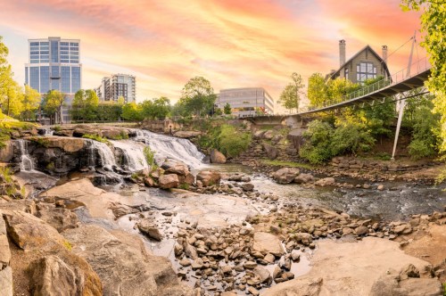 The painted pink and orange swirls of the skyline of Greenville illuminated by the setting sun watched from Falls Park on the Reedy.