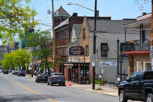 A street view of Little Italy in Cleveland, Ohio, featuring Guarino's Restaurant, a historic Italian eatery with a vintage neon sign. The scene includes brick and stone buildings, parked cars, pedestrians, lush green trees lining the street, and utility poles with overhead wires. The atmosphere reflects the charm and character of this walkable neighborhood known for its authentic Italian cuisine.