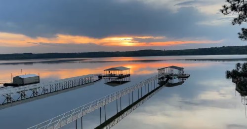 A vibrant sunset glows over a calm Mississippi lake, reflecting on the water near long piers and covered boat docks.
