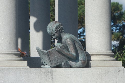 A close-up photograph of the bronze statue at the James Whitcomb Riley grave site in Crown Hill Cemetery, Indianapolis, Indiana. The statue depicts a young boy sitting cross-legged while intently reading a book titled "Riley – Poems for Children." The sculpture rests on the stone monument, surrounded by classical columns. Sunlight casts shadows on the detailed figure, highlighting its textured surface. In the background, green pine trees and part of the memorial structure are visible.