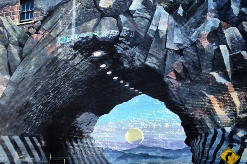 A closer view of the curvy tunnel showcases the lines on the road, the jagged rocks, and the rising sun behind the mountains.