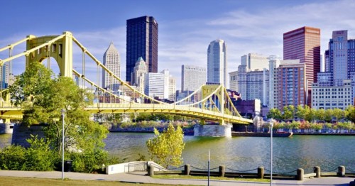 The yellow Roberto Clemente Bridge stretching across a river toward the Pittsburgh skyline under a clear blue sky.