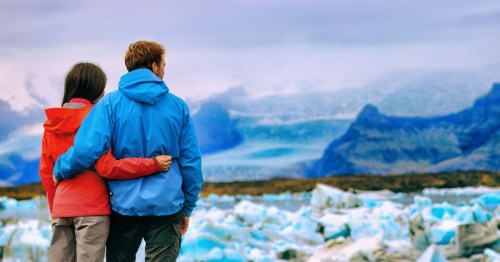 A man and woman wearing winter gear hold each other close as they look at a wintry scene on a romantic hike in Alaska.