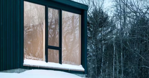 * A modern dark-paneled building features a large window reflecting a wintry forest, nestled in deep white snow.